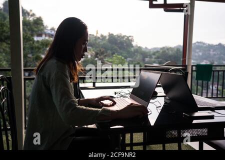 Side view of young female freelancer on vacation in casual clothing working with laptop at outdoors terrace of restaurant at Sri Lanka Stock Photo