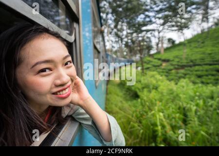 Overjoyed Asian female on vacation traveling by train along green plants while smiling and looking out window of train at Galle at Sri Lanka Stock Photo