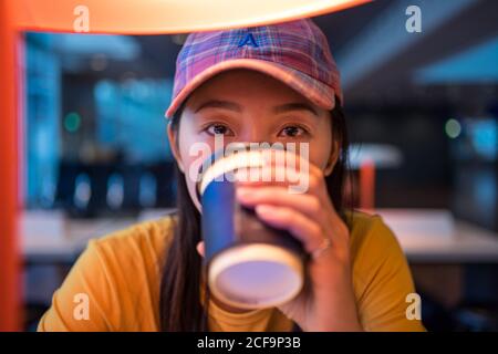 Side view of Asian Woman in cap drinking coffee from disposable cap at table with illumination and looking up in airport Stock Photo