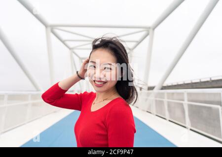 Charming Asian lady in red shirt smiling and looking at camera while walking on modern city bridge Stock Photo