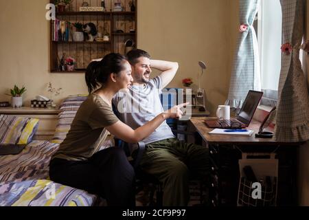 Young couple in casual clothes sitting in bedroom at table and using computer together while discussing video Stock Photo