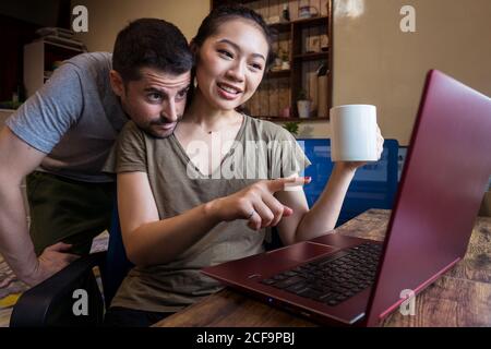 Low angle of happy boyfriend helping asian girlfriend working on the computer at home while having a hot beverage in a mug Stock Photo