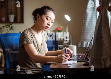 Focused female freelancer in casual clothing sitting at table with laptop and writing in notepad while working on business strategy in home office Stock Photo
