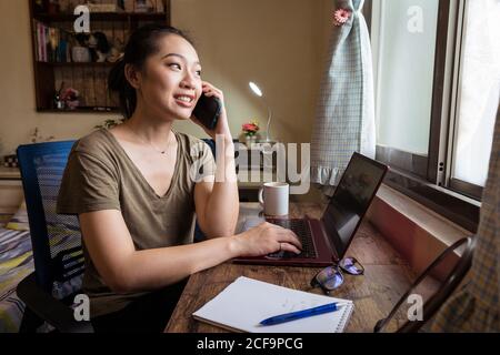 Side view of Asian female freelancer in casual t shirt sitting at table and browsing computer working on project online at home while speaking o the smartphone Stock Photo