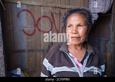 Timor Leste - August 14, 2018: High angle of calm elderly ethnic female in casual clothes looking away with interest while standing at entrance of wooden hut Stock Photo