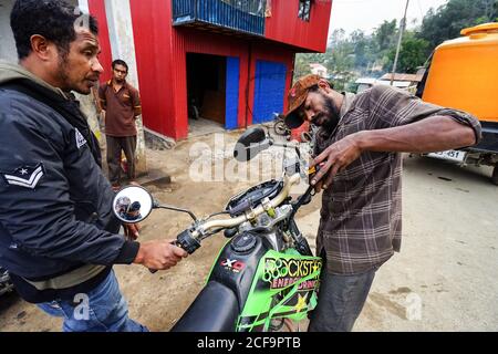 Timor Leste - August 10, 2018: Diligent ethnic mechanic carrying out maintenance of motorcycle on street standing beside client against vivid red and blue facade of garage in small town Stock Photo