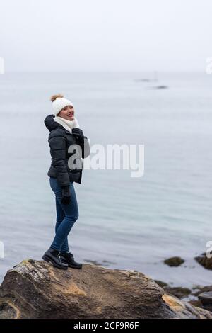 Side view of Woman in warm clothes looking at camera on small rocky ledge and enjoying beauty of northern landscape in Tromso, Norway Stock Photo