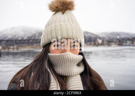 Young joyful Asian female tourist in warm clothing and hat smiling while sightseeing with mountains and sea on background at Tromso in Norwaylooking at camera Stock Photo