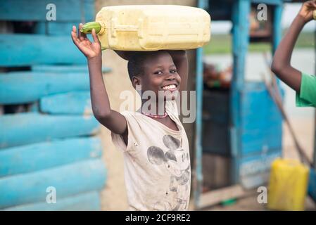 Uganda - November, 26 2016: Happy African girl smiling and looking at camera while carrying empty water canister with zucchini cork on head outside weathered house in village Stock Photo