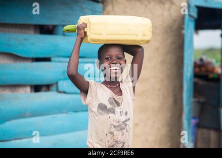 Uganda - November, 26 2016: Happy African girl smiling and looking at camera while carrying empty water canister with zucchini cork on head outside weathered house in village Stock Photo