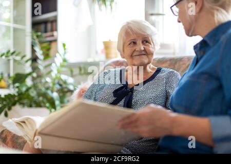 Senior woman and her adult daughter looking at photo album together on couch in living room Stock Photo