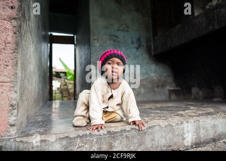 Uganda - November, 26 2016: Cute African baby in dirty pajama and knitted hat looking at camera while sitting inside uncompleted concrete building Stock Photo