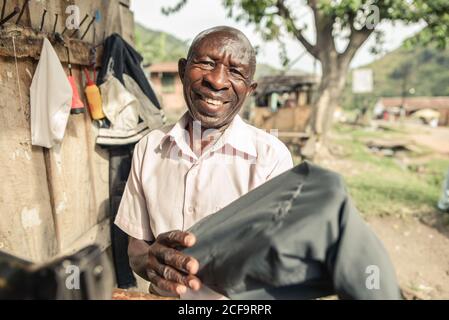 Uganda - October, 15 2016: Happy black man smiling and looking at camera while showing off a piece of garment sewed on machine outside shabby workshop on street Stock Photo