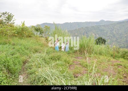 Uganda - November, 26 2016: African woman mother carrying adorable baby in sling with two other kids looking at camera while walking in bushy mountain in countryside Stock Photo