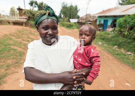 Uganda - November, 26 2016: Friendly African woman with cute girl smiling and looking at camera while standing outside house on village street Stock Photo