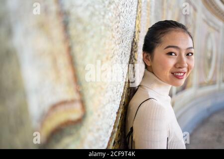 Side view of content Asian female on vacation in beige roll neck leaning on wall with fresco while smiling and looking at camera at St Peters Basilica in Vatican at Rome Stock Photo