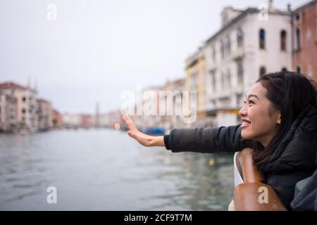 Side view of overjoyed Asian resting lady in warm clothing smiling and reaching out from boat on waterway amid old buildings