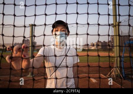 Young ethnic female in sportswear and protective mask for coronavirus prevention looking at camera through net fence while standing on empty sports ground in city
