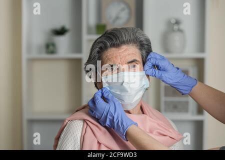 Crop anonymous social worker in latex gloves putting on medical mask on elderly female on chair while taking care of pensioner during coronavirus pandemic at home