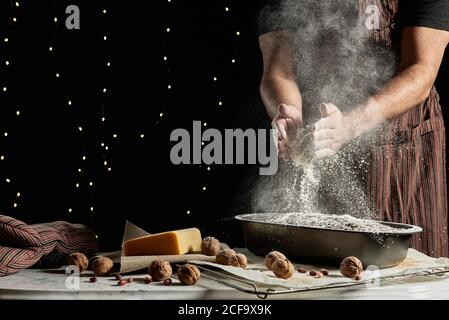 Crop anonymous male baker in apron spilling flour over baking pan with bread while working at table with ingredients Stock Photo