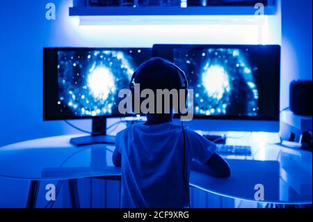 Back view of unrecognizable male kid in headphones watching film about planets on desktop computer while sitting at table in room with blue light Stock Photo