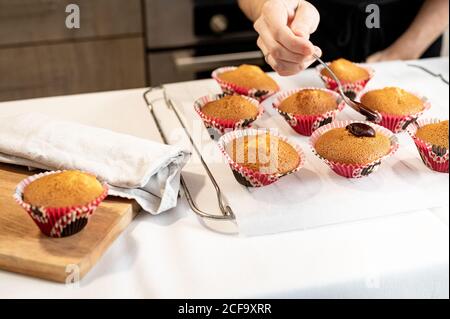 Crop Woman using spoon to spread chocolate frosting on freshly baked cupcakes while preparing pastry at home Stock Photo