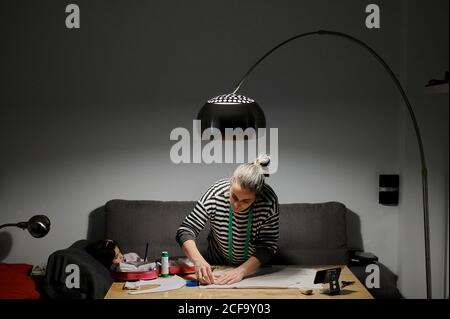 Focused female designer in casual clothes working with fabric in light of modern lamp at wooden table near sofa at home Stock Photo