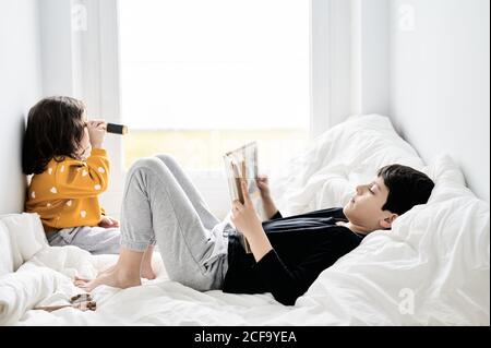 Side view of brother reading book and sister playing with retro spyglass relaxing on comfortable bed and enjoying weekend Stock Photo
