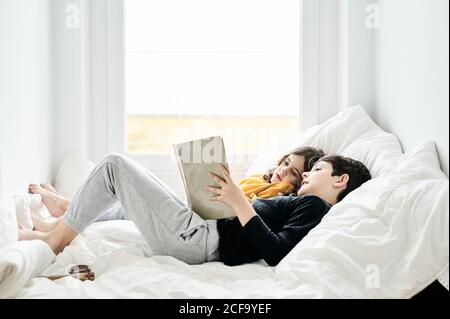 Side view of relaxed brother and sister lying on soft bed and reading fairy tale together while enjoying weekend at home Stock Photo