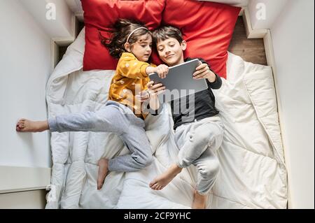 Top view of brother and sister lying on soft blanket and pillows and watching cartoon on tablet while entertaining during weekend Stock Photo