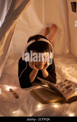 Thoughtful male child in headset reading book with images while listening to music and lying in cozy kids play tent in apartment Stock Photo