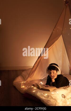 Thoughtful male child in headset reading book with images while listening to music and lying in cozy kids play tent in apartment Stock Photo