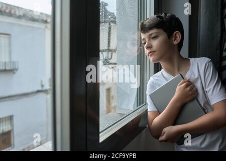 Thoughtful child in casual t shirt standing with tablet and leaning on window while observing rain in street Stock Photo