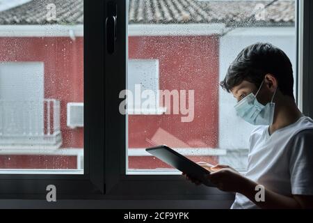 Side view of kid in casual wear browsing tablet while standing near window during quarantine Stock Photo