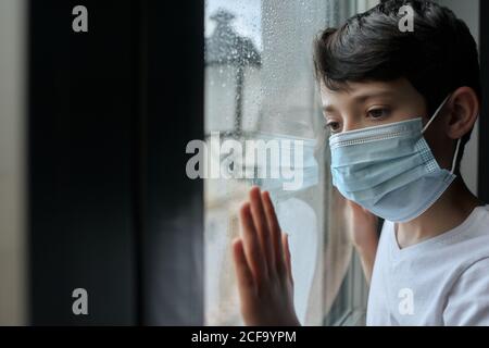 Unhappy child wearing face mask standing near window and observing street while staying home during coronavirus epidemic Stock Photo