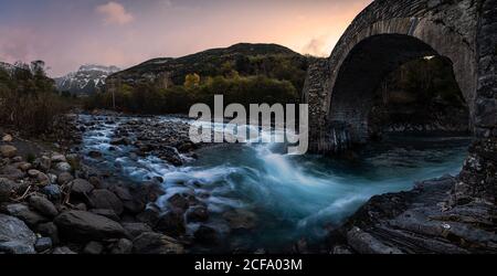 Panoramic view of old stone arched bridge crossing mountain river flowing through rocky valley in forest during sunset in autumn Stock Photo