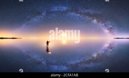 Silhouette of anonymous man standing on reflection surface of water and reaching out to starry colorful night sky with milky way Stock Photo