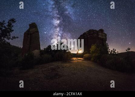 Silhouette of man with torch in hand standing on hill with big ricks among green plants with colorful night sky with stars and milky way on background Stock Photo