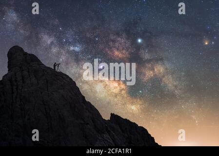 From below of silhouette of traveler standing on rough rock and looking through telescope while enjoying stunning view of amazing starry sky with breaking rays of rising sun Stock Photo