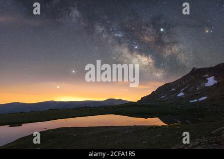 Amazing picturesque landscape of rising sun from behind snowy mountain range against background of night sky with star lighting and reflecting in clear mountain lake on winter night Stock Photo