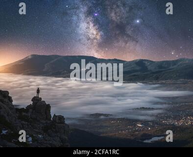 Drone view of traveler watching sunrise while standing on cliff and enjoying stunning views of majestic mountains above clouds spreading out at foot of mountains and amazing starry sky Stock Photo