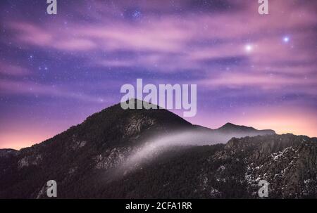 Majestic view of cloudy purple starry sky with rising sunbeams from behind snowy mountain range on cold winter beautiful night Stock Photo