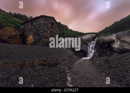 Low angle of picturesque view of rocky terrain with old stone building located near water flow and surrounded by mountains Stock Photo