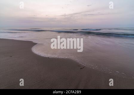 Sea waves rolling on wet sandy coast against cloudy sky in evening in nature Stock Photo