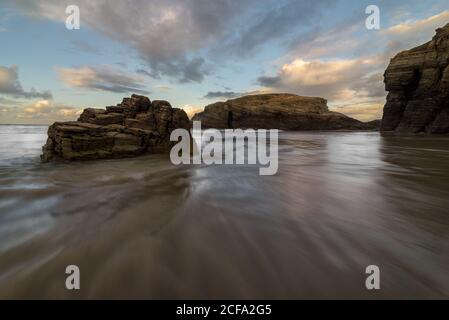 Big rocky formations on sandy empty beach of ocean with cloudy bright evening sky on background Stock Photo