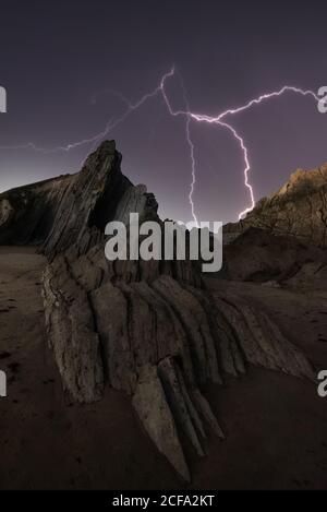 From below rough dramatic landscape of rocky cliff under dark stormy sky with bright lightnings at night Stock Photo