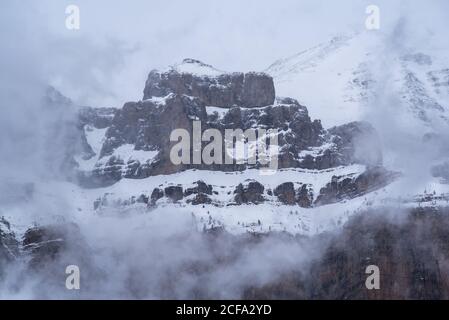 Sharp mountain peaks covered partially with snow surrounded by misty fog under cloudy sky in wintertime Stock Photo