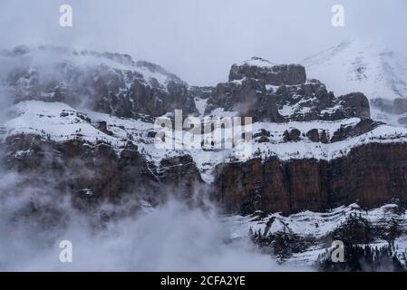 Sharp mountain peaks covered partially with snow surrounded by misty fog under cloudy sky in wintertime Stock Photo