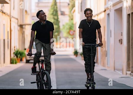 Cheerful young African American man looking at camera and riding electric scooter while black male friend looking up and driving bicycle in street Stock Photo
