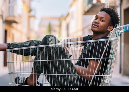 Side view of pensive youthful African American male teenager in casual clothes looking at camera while sitting in metal shopping trolley in street Stock Photo